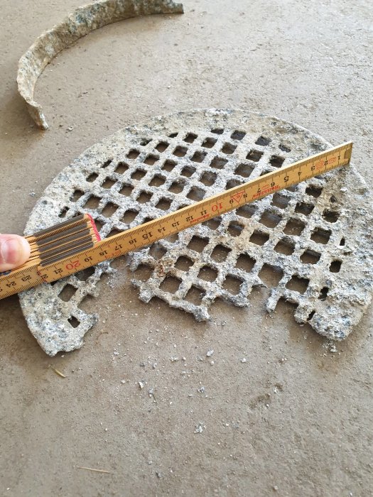 Slitet avloppsgaller av metall med ett måttband som visar diametern på 240mm och en trasig distansring på en garagegolv.