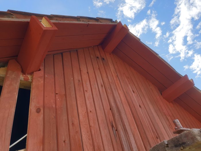 En nyförlängd och upprättad takbjälke mot en ojämn röd trävägg under blå himmel.