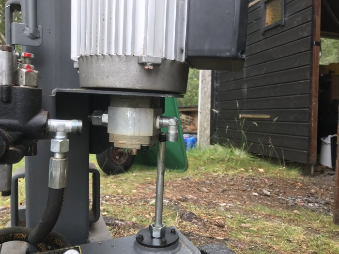 Hydraulmotor och pump från en vedklyv, ihopkopplade och monterade på en metallplatta, med verktygsskjul i bakgrunden.