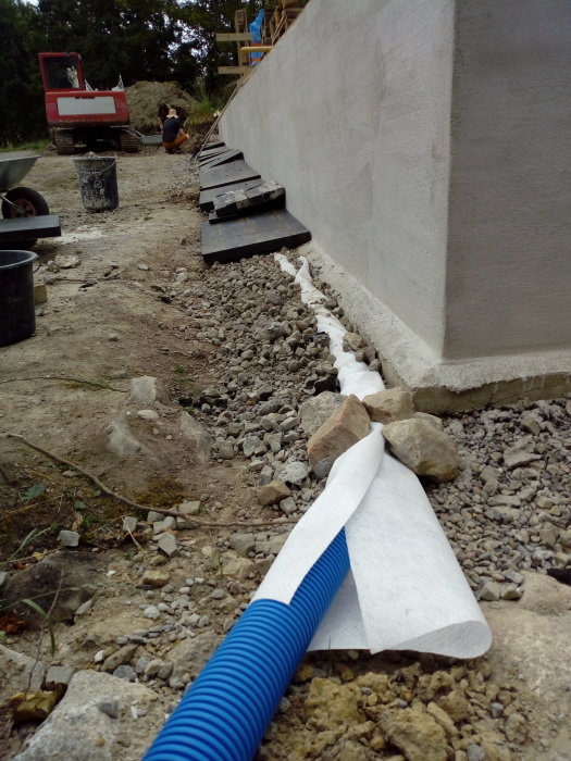 Dräneringsrör och markisolering längs husgrund med byggmaterial och grävmaskin i bakgrunden.