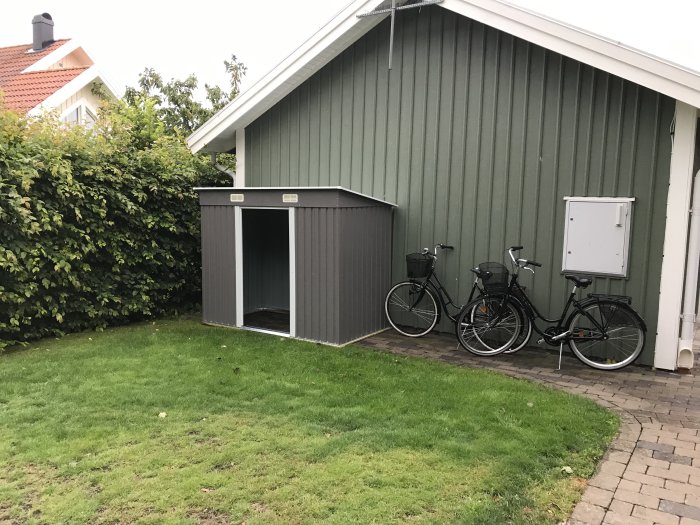 Ett litet grått trädgårdsförråd utan dörrar står på en lutande stenläggning bredvid en grönskande häck och två parkerade cyklar.