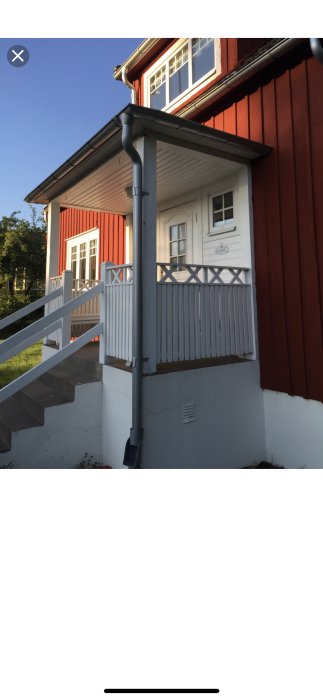 Hörnet av ett rött trähus med vit veranda och trappa i behov av renovering under en klarblå himmel.