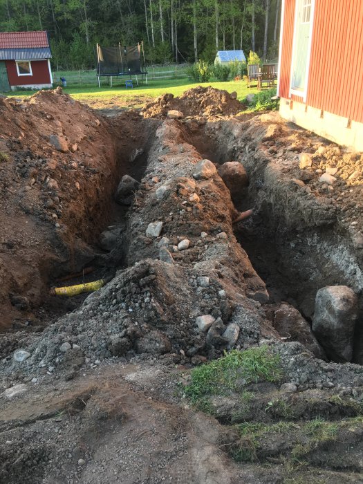 Grävd ränna med stenar och en gul PVC-rör vid en byggplats nära ett hus.