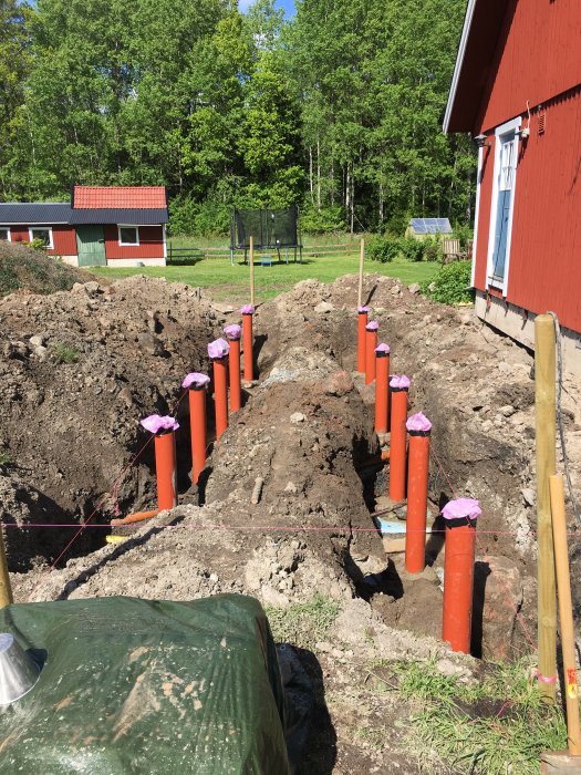 Gjutna plintfötter i en grävd ränna med PVC-rör, i stenig jord nära en röd byggnad.