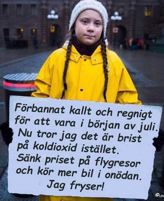 Barn i gul regnjacka och vit mössa håller ett skylt med text om kallt väder och en ironisk kommentar om koldioxid och bilkörning.