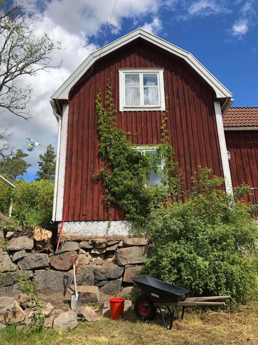 Hörn av gammalt hus med skadad stenmur och verktyg för trädgårdsarbete, stubbe och grönska syns.