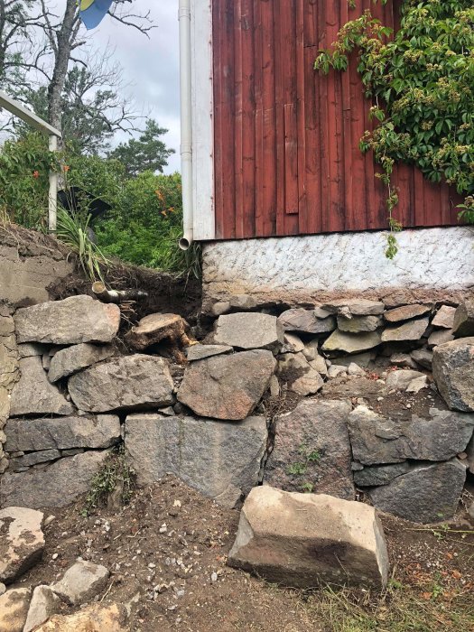 Hörn av gammal naturstensmur med synliga skador och trädrot efter stubbavlägsning vid rödfärgat hus.