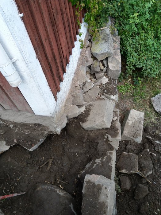 Hörnet av ett gammalt hus med en delvis nedmonterad naturstensmur och synliga rötter i marken.