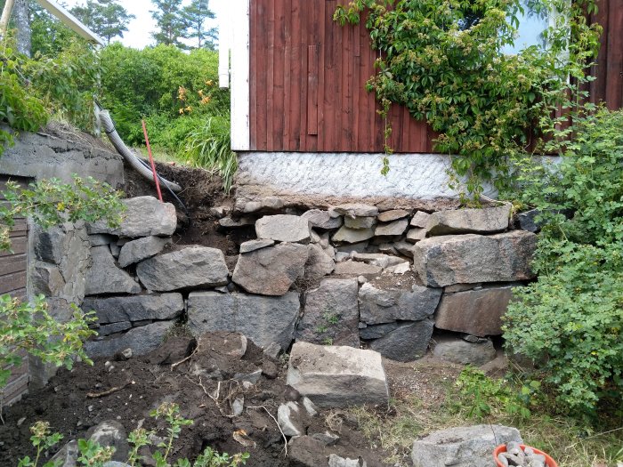 Hörnet av ett gammalt hus med en delvis raserad naturstensmur och synliga jordhögar efter borttagningen av ett träd.