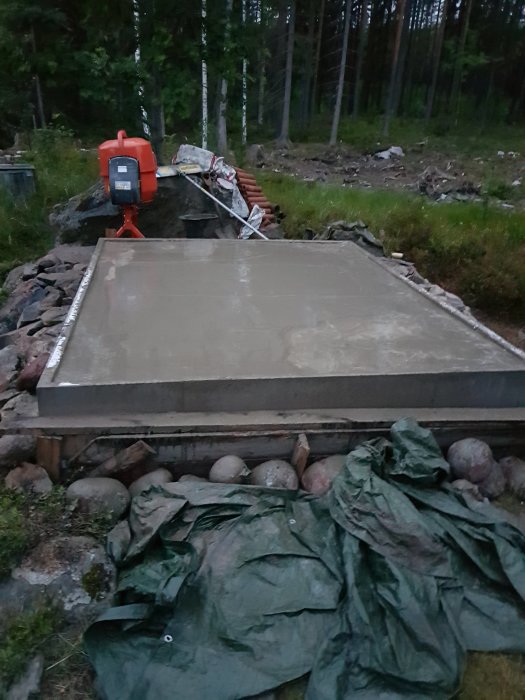 Nygjuten betongplatta i dagsljus med synlig kant där cement saknas, omgiven av skogsmark och byggmaterial.