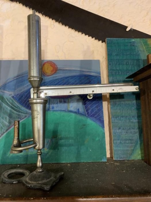 Gammaldags höjdjusterbar lamphållare med metallarm och justeringsskruv på ett bord framför abstrakt konst.
