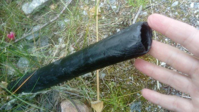 Hand håller ett avgrävt, svart rör vid husknuten med gräs i bakgrunden.