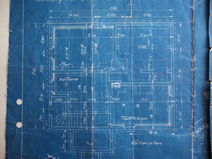 Blått arkitekturritning av ett källarplan med måttangivelser och rumsmarkeringar på slitet papper.