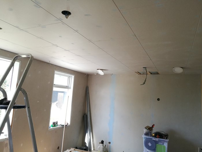 Renoveringsarbete i rum med oslipad spackel på gipsskivor i tak och längs en vägg, en stege och verktyg syns.