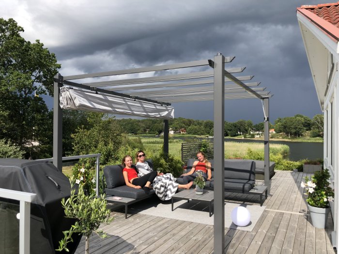 Nybyggd pergola med sittgrupp på trädäck, mörka moln i himlen, personer kopplar av utomhus.