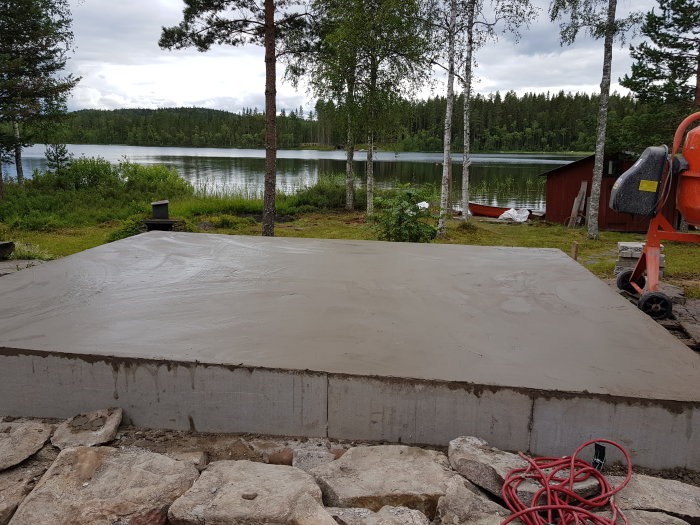Nyutgjuten ojämn betongplatta med betongblandare i hörnet nära en sjö och röd stuga.