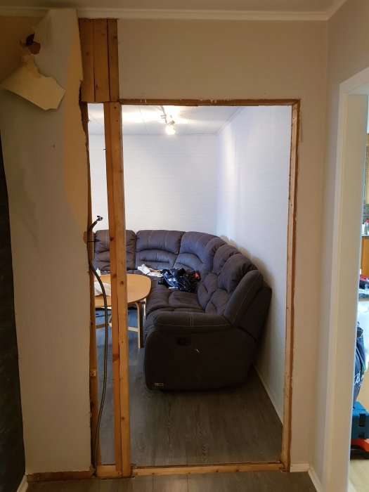 En nedkapad dörrkarm i en vägg med en synlig tjock trästolpe, utsikt mot ett vardagsrum med soffa och bord.