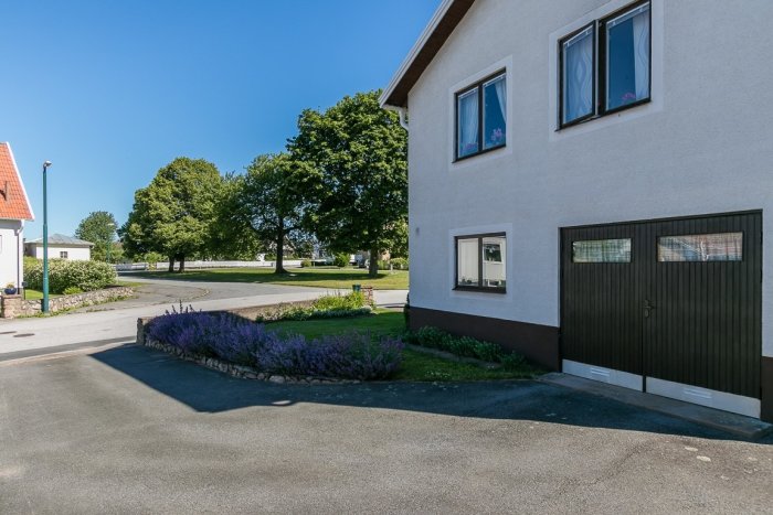 Husens entré utan synlig gångväg, asfalterad uppfart, lavendelrabatt, garageport och gräsmatta.