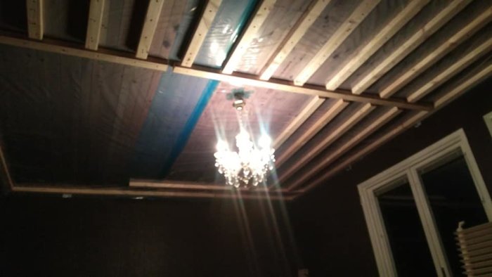 Unfinished wooden ceiling beams with insulation and a crystal chandelier in a renovation project.