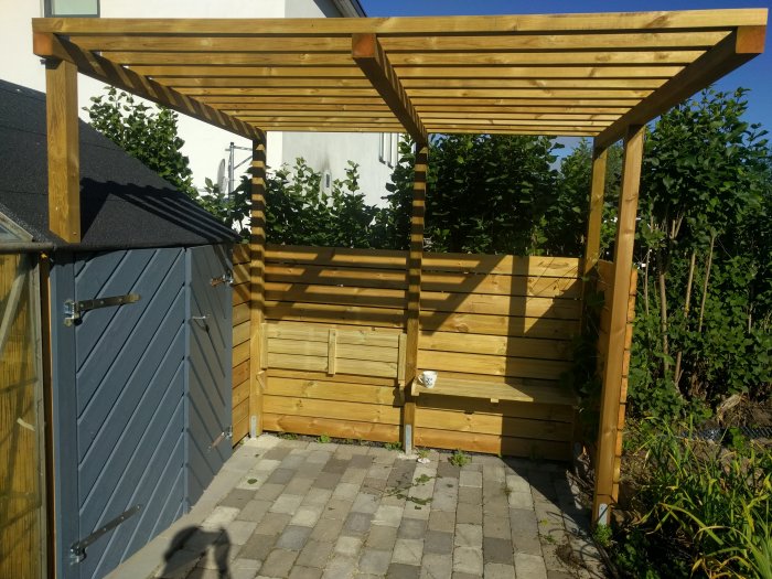 Nybyggd pergola med sittbänkar och dold lucka vid ett förråd, omgiven av grönska.