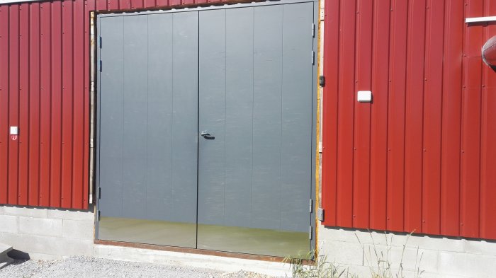 Gråmålade dubbla dörrar av plywood med synliga sprickor i färgen och skador från väder och vind.