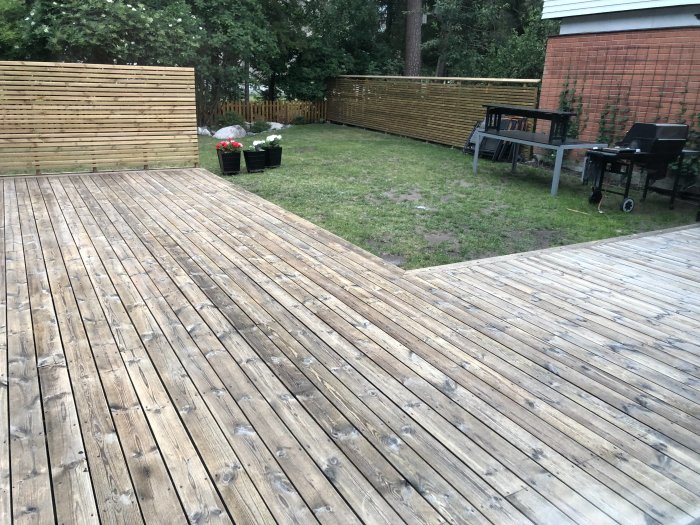 Trädäck i en trädgård med gräsmatta, trädgårdsmöbler och grill intill en tegelbyggnad.