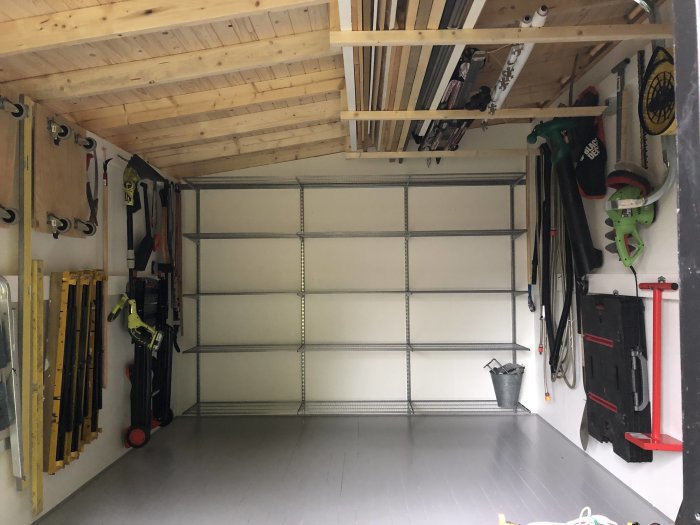 Ett välordnat och rent garage med verktyg upphängda på väggen, träbjälkar i taket och en stängd garageport.