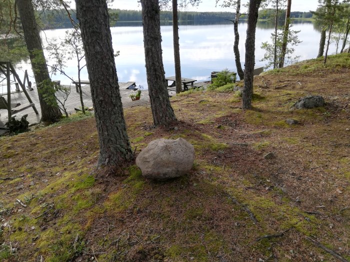 Stor bojsten på gräsmark med träd i förgrunden och brygga och sjö i bakgrunden.