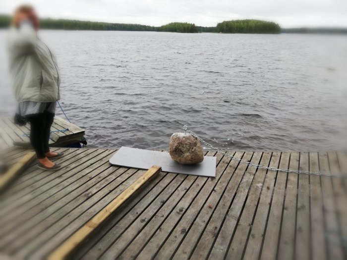 Bojsten med lyftögla och kätting på en brygga vid sjö, person delvis synlig, molnig dag.