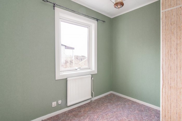 Grönmålad rumshörna i gammalt hus med fönster, radiator, och träpanelvägg redo för renovering.