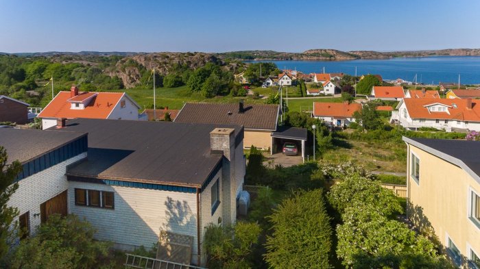 Flygbild av ett gammalt sommarhus med papptak omgivet av grönska i Bohuslänsk fiskeby, havsutsikt i bakgrunden.