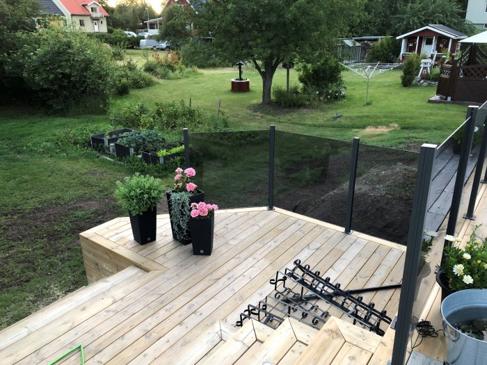 Upphöjd träterrass med ofärdigt glasräcke, blomkrukor och en trädgård i bakgrunden.