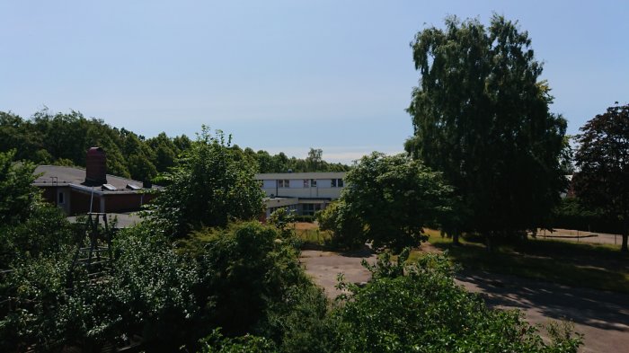 Utsikt från balkong med buskar i förgrunden och vitt tvåvåningshus i plåt i bakgrunden under en klar himmel.