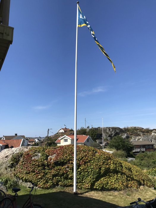 Flaggstång med blå och gul flagga i en trädgård med röda hus och grönska i bakgrunden.