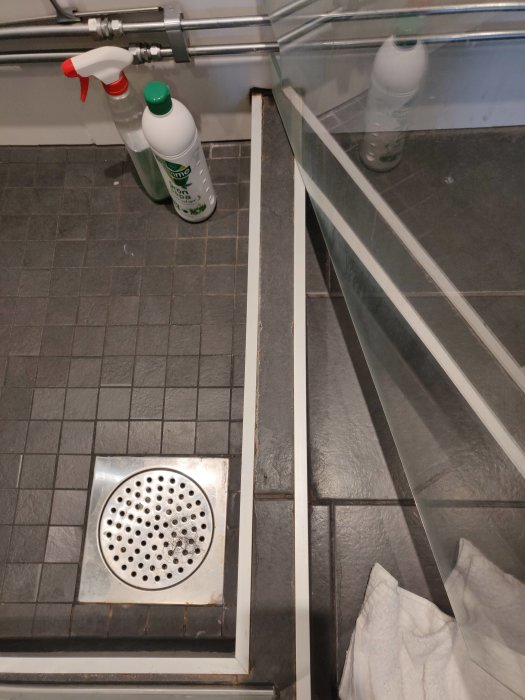Badrumsgolv med en duschutrymmets kant och glasvägg, problem med vattenavrinning närmast metallisten.