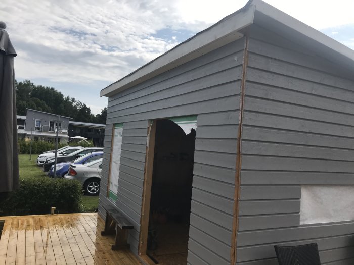 Halvfärdigt bygge av ett litet gråmålat trädhus med takstolar och grundmålade brädor, under en molnig himmel.