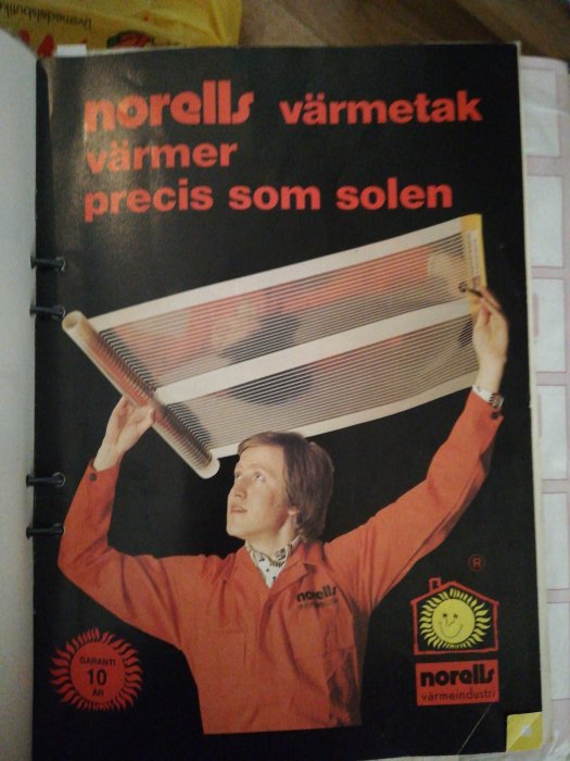 Person i orange jacka håller en värmeelementpanel ovanför huvudet mot en svart bakgrund med texten "värmer precis som solen".