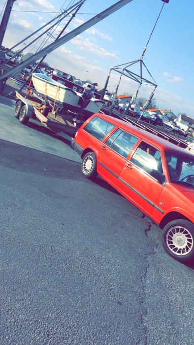 Röd bil med båt på trailer i en hamn, med andra båtar och kran i bakgrunden.