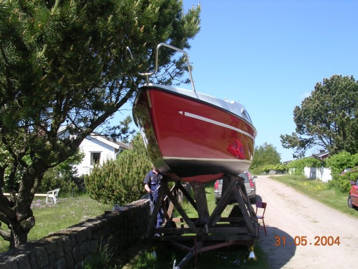 Röd segelbåt på en obromsad trailer parkerad på en landväg omgiven av gröna träd och hus.