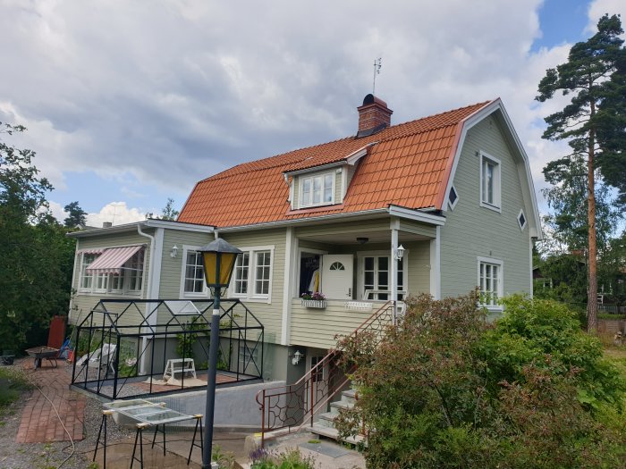 Grönmålat hus med tegeltak, veranda och trädgård, teglens färg mörknar med åldern.