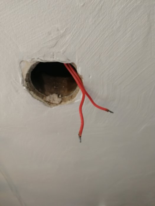 Uppstickande röda elkablar ur ett runt hål i en vit vägg.