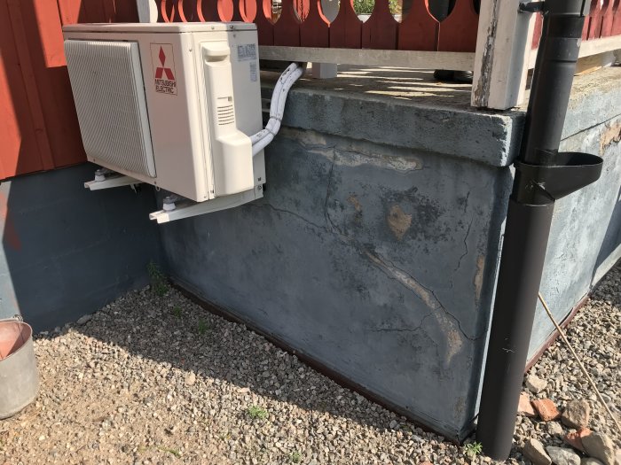 Luftvärmepump installerad på en vägg ovanför ett grusområde, med synligt skadade betongstenar och platonmatta vid husgrund.