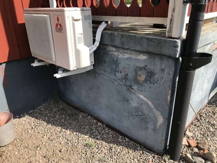 Utsidan av en husgrund med synligt skadad betong och en installerad luftvärmepump ovanför grussäng.