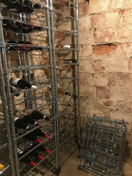 Ett potentiellt vinkällarutrymme med tegelväggar och metallvinhyllor fulla med flaskor, några skadade tegel syns.