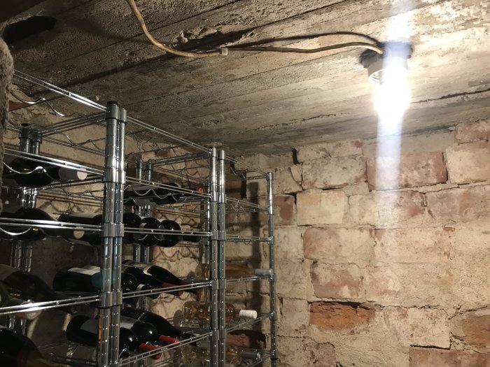 Vinkällare med skadat tegel på vägg, metalliskt vinhyllsystem, synlig belysning och betongtak som visar slitage och saltutfällningar.