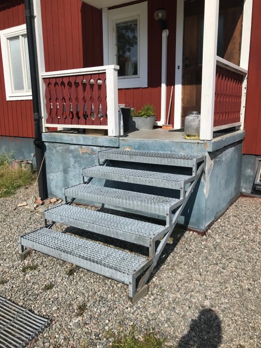 Trappsteg av metall leder upp till en entré under rödmålad husfasad med betonggrund, möjlig ingång till vinkällaren.