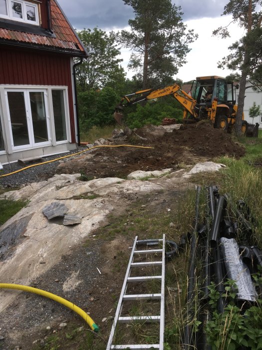 Traktorgrävare gräver vid husrad för framtida altan, stora stenar och jordhögar synliga.