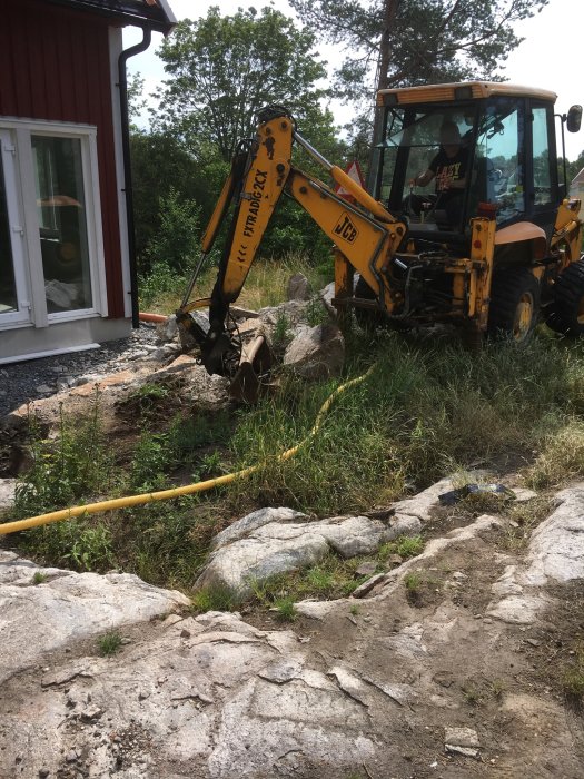 Liten traktorgrävare som gräver bland stenar nära en röd husvägg, delvis molnig himmel.