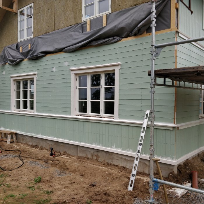 Hus under renovering med grön panelbräder, kärnfura vattbräda ovan fönster, byggställning och stege.