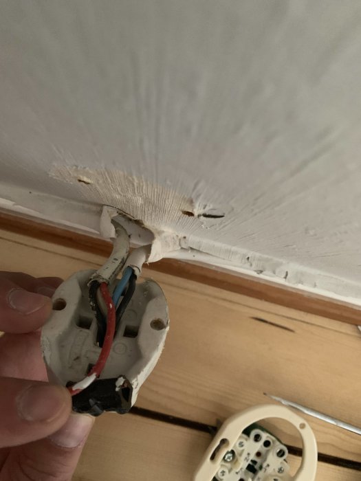 Hand som håller en demonterad elektrisk sockel med synliga ledningar under ett ojämnt tak.